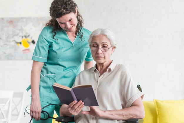 Enfermera sonriente que mira el asimiento del libro por el paciente mayor femenino que se sienta en la silla de rueda