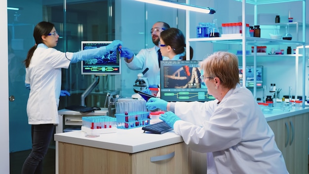 Enfermera sosteniendo tablet PC con información científica mientras químico usando microscopio con tubo de ensayo químico cerca