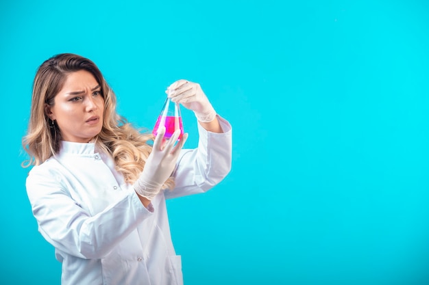 Enfermera en uniforme blanco sosteniendo un matraz químico con líquido rosa y parece dudosa.