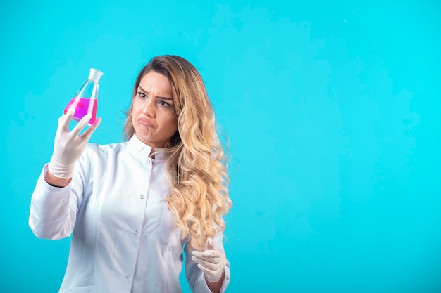 Enfermera en uniforme blanco sosteniendo un matraz químico con líquido rosa y parece dudosa.