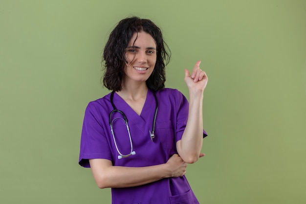 Enfermera con uniforme y un estetoscopio apuntando hacia arriba y mirando a la cámara con una sonrisa en la cara de pie en verde aislado