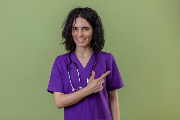 Enfermera con uniforme y un estetoscopio apuntando hacia el lado con el dedo índice con una sonrisa en la cara de pie en verde aislado