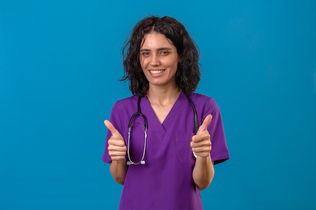 Enfermera con uniforme y estetoscopio con sonrisa de confianza mostrando los pulgares hacia arriba con cara feliz de pie en azul aislado