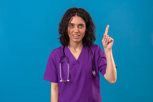 Enfermera vestida con uniforme y un estetoscopio apuntando hacia arriba con el dedo se recuerda a sí misma para no olvidar lo importante de pie en azul aislado