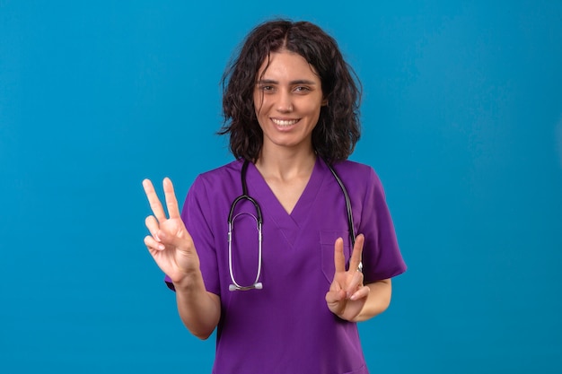 Enfermera vistiendo uniforme y estetoscopio sonriendo alegre mirando a un lado y mostrando el número dos o signos de victoria con los dedos en azul aislado