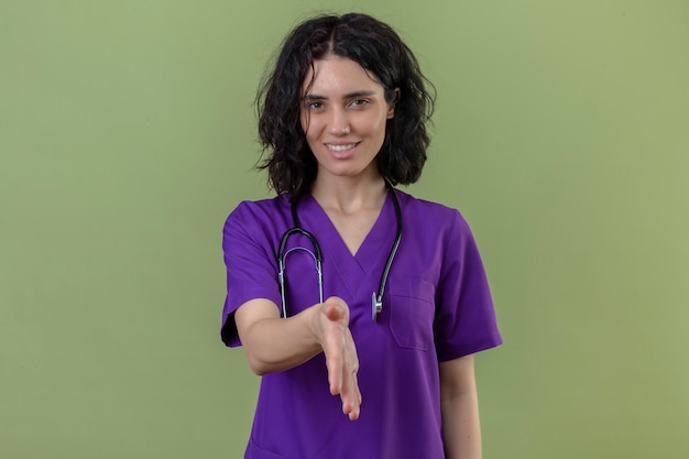Enfermera vistiendo uniforme y estetoscopio sonriendo amable ofreciendo mano haciendo gesto de saludo en verde aislado