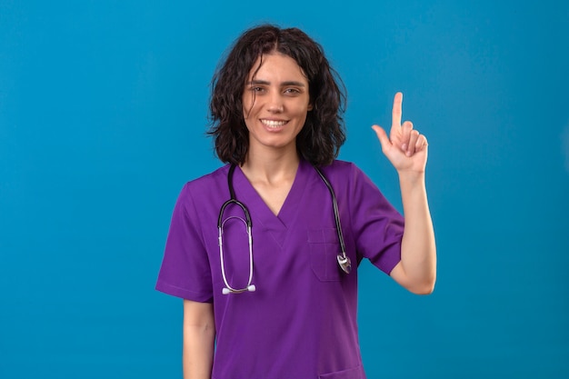 Enfermera vistiendo uniforme médico y con estetoscopio apuntando hacia arriba con el dedo sonriendo confiado concentrado en la tarea de pie en azul aislado