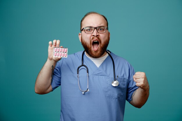 Enfermero joven enojado con anteojos exfoliante de enfermera y estetoscopio alrededor de su cuello mostrando un paquete de pastillas y el puño mirando a la cámara gritando aislado sobre fondo azul.