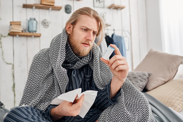 Enfermo hombre rubio con barba en ropa de dormir se sienta en la cama rodeado de mantas y almohadas, frunce el ceño mientras lee la receta de las píldoras y sostiene un pañuelo en la mano. Problemas de salud, resfriado y gripe.