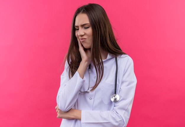 Enfermo joven médico con estetoscopio bata médica puso su mano sobre el dolor de muelas sobre fondo rosa aislado