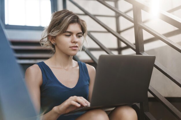 Enfocada, guapa, rubia, joven de 20 años, mujer independiente, trabajando en un nuevo proyecto, sola, al aire libre, sentarse, escaleras, sostener las rodillas de la computadora portátil, mirar determinada, netbook, dispositivo, pantalla, teclado, estudiar antes de los exámenes finales.