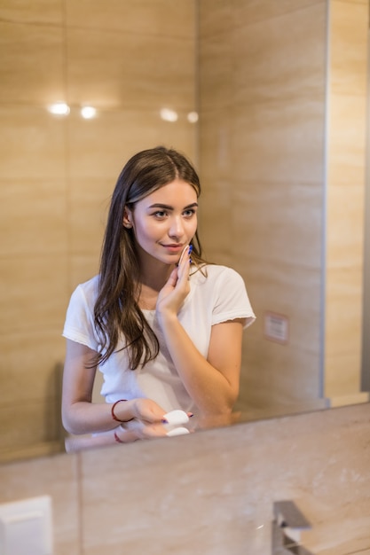 Enfocada hermosa joven limpiando su piel con un algodón, mirando el espejo en el baño de su casa