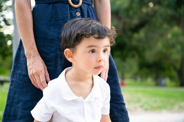 Enfocado emocionado niño de pelo negro de pie junto a mamá al aire libre y mirando a otro lado. Tiro medio. Concepto de infancia