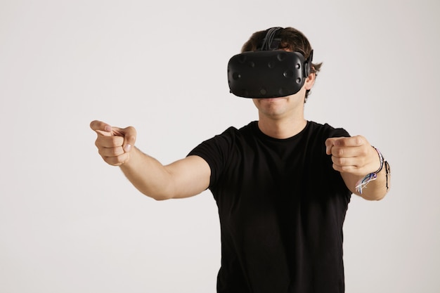 Enfocado joven jugador serio en camiseta negra y gafas VR estirando las manos como si condujera, aislado en blanco
