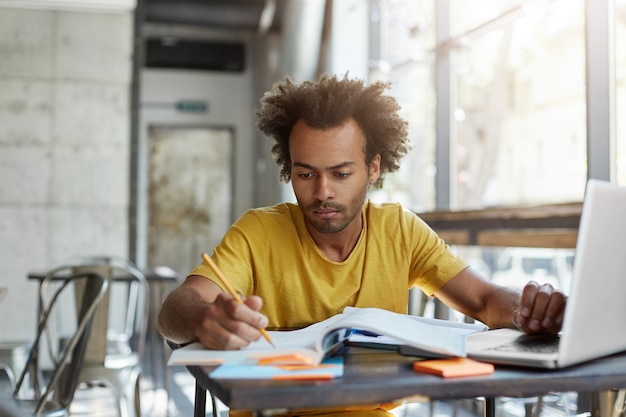 Enfocado joven profesor de inglés afroamericano revisando los cuadernos de sus estudiantes, sentado en la mesa de café frente a la computadora portátil abierta. Grave lección de aprendizaje de estudiante masculino negro en el comedor universitario