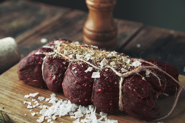 Enfoque cercano cuerda atado trozo de carne salada con pimienta lista para fumar en la mesa de madera entre hierbas