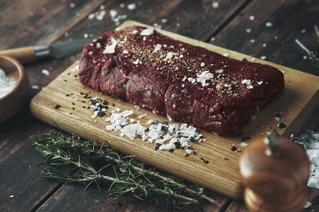 Enfoque cercano trozo de carne salada con pimienta lista para asar en la mesa de madera entre hierbas