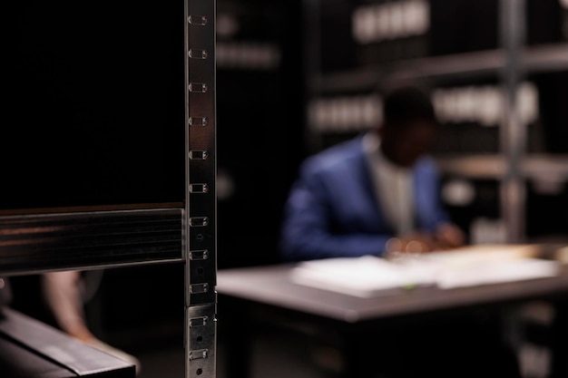 Foto gratuita enfoque selectivo de estantes llenos de documentos policiales, en segundo plano investigador analizando pruebas de la escena del crimen. detective privado afroamericano trabajando en un caso de persona desaparecida en una sala de archivo.