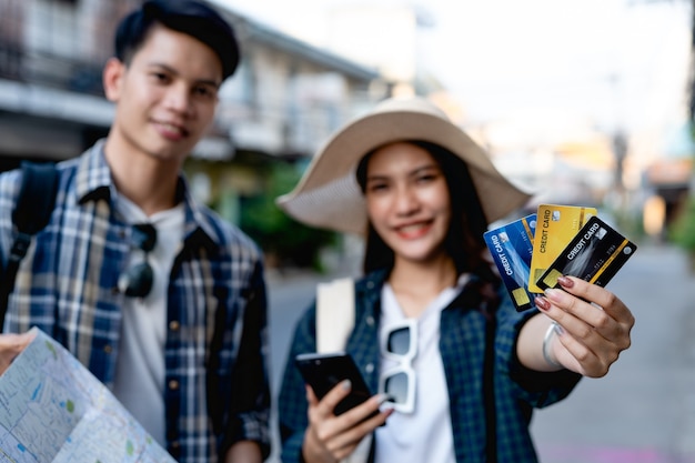 Enfoque selectivo, joven mochilero sosteniendo un mapa de papel y una mujer bonita con sombrero sostiene un teléfono inteligente y muestra una tarjeta de crédito en la mano.Los usan para pagar el viaje con felicidad en vacaciones.