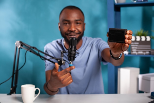 Foto gratuita enfoque selectivo en la mano de vlogger que presenta la revisión del paquete de baterías recargables npf que señala las características del producto sentado en el escritorio. influencer revisando un acumulador de iones de litio usando un micrófono profesional