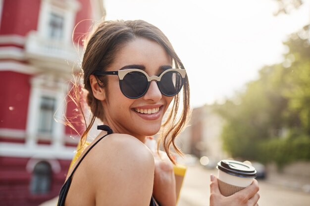 Enfoque suave. Concepto de estilo de vida. Ciérrese encima del retrato de la mujer caucásica morena joven atractiva alegre alegre en gafas de sol y traje negro que sonríe con los dientes, tomando café, sosteniendo bolsas con ropa af