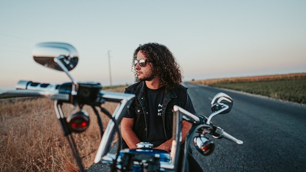 Enfoque superficial de un hombre fresco de pelo rizado con una chaqueta de mezclilla negra en su motocicleta en la carretera
