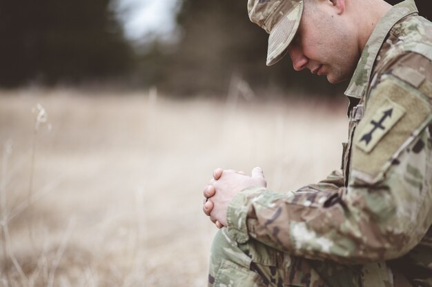 Enfoque superficial de un joven soldado rezando mientras está de rodillas sobre una hierba seca