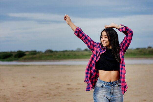 Enfoque superficial de una mujer hispana alegre adulta vistiendo un top corto con una camisa de franela en una playa