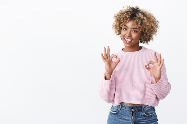 Enfriar todo perfecto. Retrato de cliente afroamericano feliz y encantado que recomienda un producto de buena calidad que muestra un gesto aceptable y una sonrisa satisfecha sobre la pared blanca