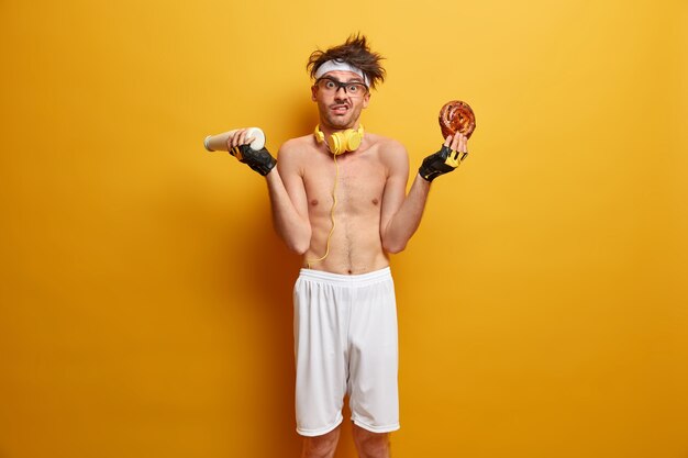 Engañar comida, calorías y concepto de deporte. Deportista vacilante con el torso desnudo, se siente desconcertado, lucha contra la tentación de comer comida chatarra, sostiene la leche y el pan, ansioso por estar en forma y deportivo, usa pantalones cortos, diadema