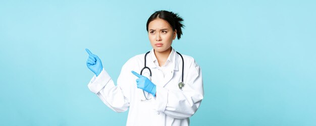 Enojado asiático trabajador médico enfermera o médico expresa desaprobación apuntando a la izquierda y surcando las cejas sta