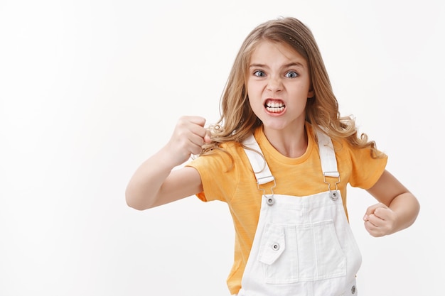 Enojado, disgustado y angustiado odiosa niña rubia, niño mirando furioso y molesto, quejándose, haciendo muecas molesta intensa pelea, mirada enojada gesticulando, apretando el puño, parado pared blanca