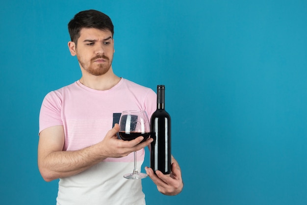 Enojado guapo y sosteniendo una copa de vino y una botella en un azul.