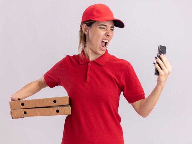 Enojado joven bonita repartidora en uniforme tiene cajas de pizza y mira el teléfono en blanco