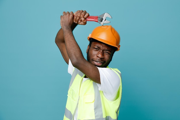 Enojado joven constructor afroamericano en uniforme con llave de gas aislado sobre fondo azul.
