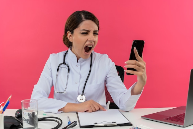 Enojado joven doctora vistiendo bata médica y estetoscopio sentado en el escritorio con herramientas médicas y portátil sosteniendo y mirando el teléfono móvil