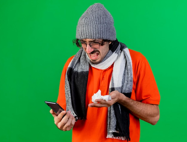 Enojado joven enfermo con gafas, gorro y bufanda de invierno sosteniendo y mirando el teléfono móvil y sosteniendo la servilleta aislada en la pared verde con espacio de copia