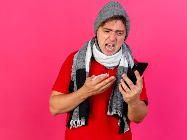 Enojado joven guapo rubio enfermo con sombrero de invierno y bufanda sosteniendo teléfono móvil y servilleta mirando teléfono aislado en pared rosa con espacio de copia