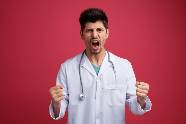 Enojado joven médico masculino vistiendo uniforme médico y estetoscopio alrededor de su cuello mirando a la cámara manteniendo los puños en el aire aislado sobre fondo rojo.