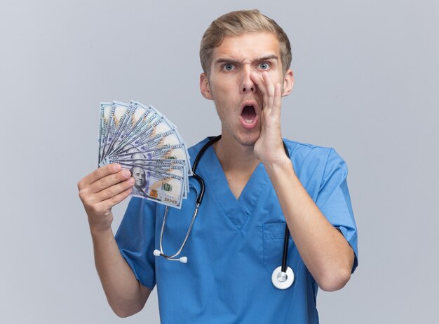 Enojado joven médico vistiendo uniforme médico con estetoscopio sosteniendo dinero en efectivo llamando a alguien aislado en la pared blanca