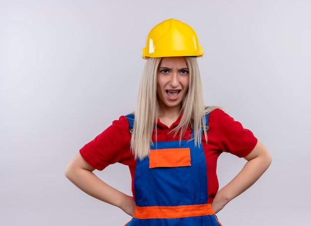 Enojado joven rubia ingeniero constructor chica en uniforme y aparatos dentales con las manos en la cintura en el espacio en blanco aislado