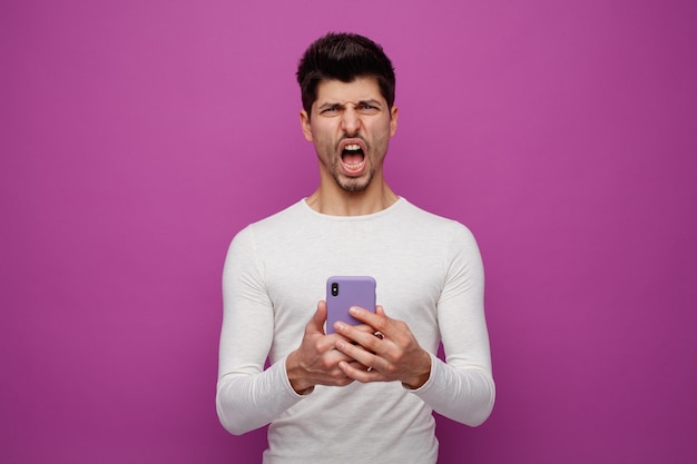 Enojado joven sosteniendo teléfono móvil mirando a la cámara gritando aislado sobre fondo púrpura