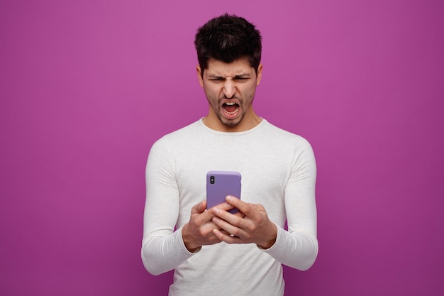 Enojado joven usando su teléfono móvil gritando aislado sobre fondo púrpura