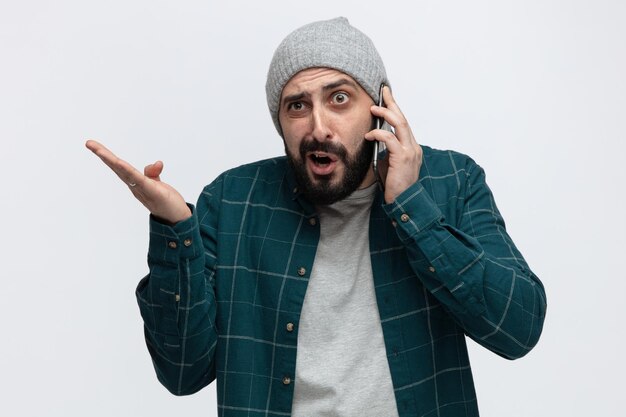 Enojado joven vistiendo sombrero de invierno mirando al lado hablando por teléfono mostrando la mano vacía aislado sobre fondo blanco.