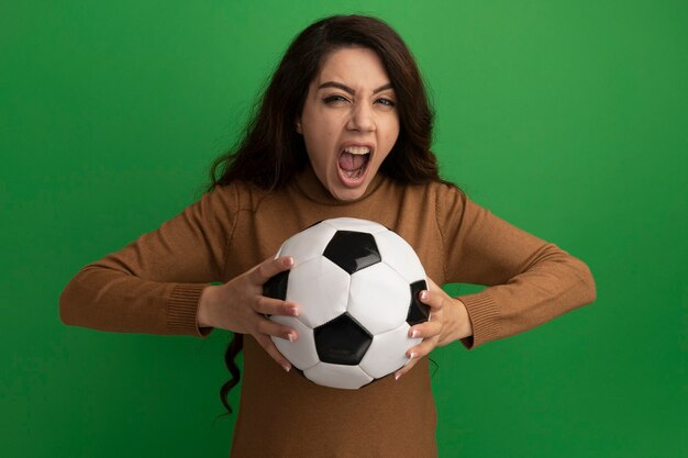 Enojado mirando a cámara hermosa joven sosteniendo la bola aislada en la pared verde