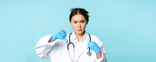 Enojado mujer asiática médico médico en uniforme y guantes muestra los pulgares hacia abajo surco cejas malestar dis