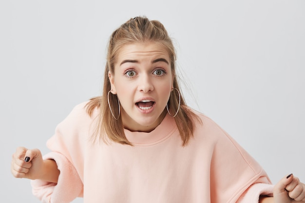 Enojado mujer joven caucásica con piel limpia sana vistiendo suéter rosa posando aislado, haciendo estallar sus ojos, listos para discutir. Emociones negativas, insatisfacción, agresión.