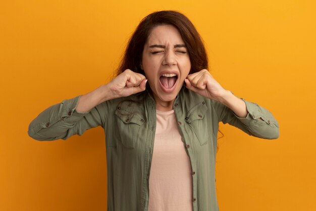 Enojado con los ojos cerrados hermosa joven vestida con camiseta verde oliva sosteniendo el puño alrededor de la cara aislada en la pared amarilla