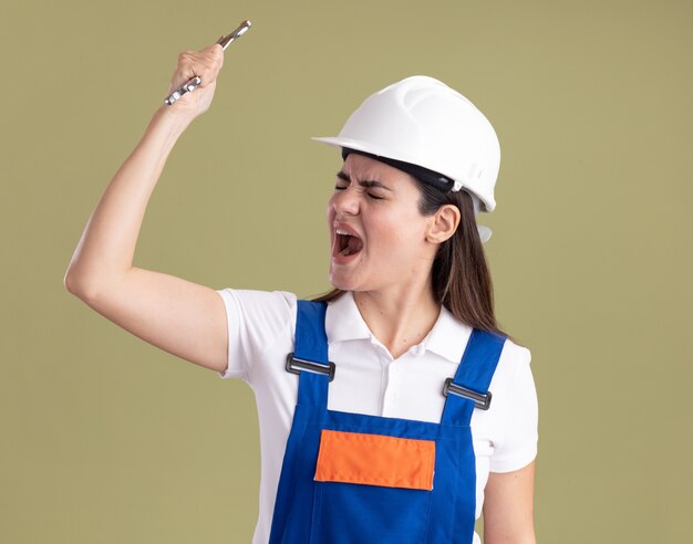 Enojado con los ojos cerrados joven constructor mujer en uniforme sosteniendo una llave de boca aislada en la pared verde oliva