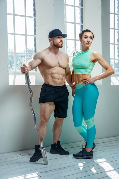 Enorme culturista barbudo sin camisa con una mujer deportiva con ropa deportiva azul posando con luz natural cerca de la ventana.